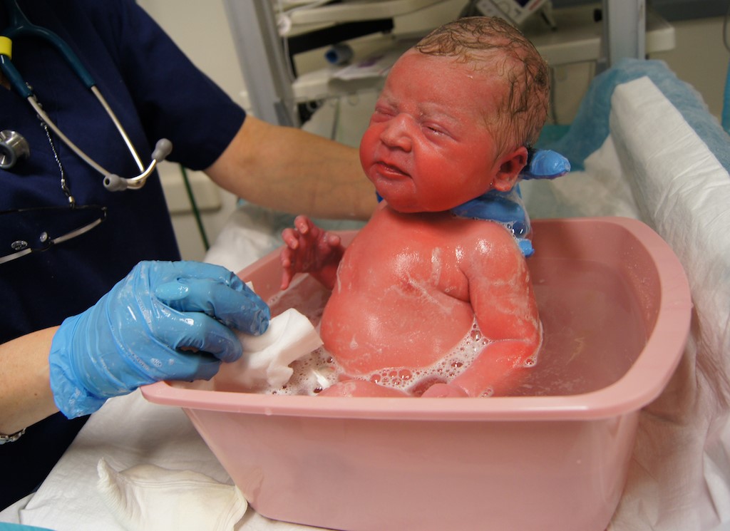 Gambar bayi sedang dimandikan oleh ibu bapa dengan berhati-hati menggunakan tuala dan air suam.