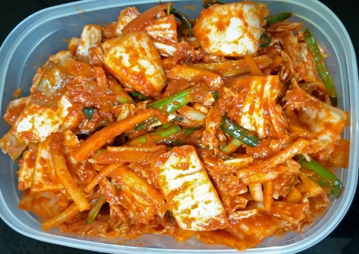 Kimchi buatan sendiri dalam bekas kuning dengan imej lobak merah, lobak putih dan daun bawang di sekelilingnya.