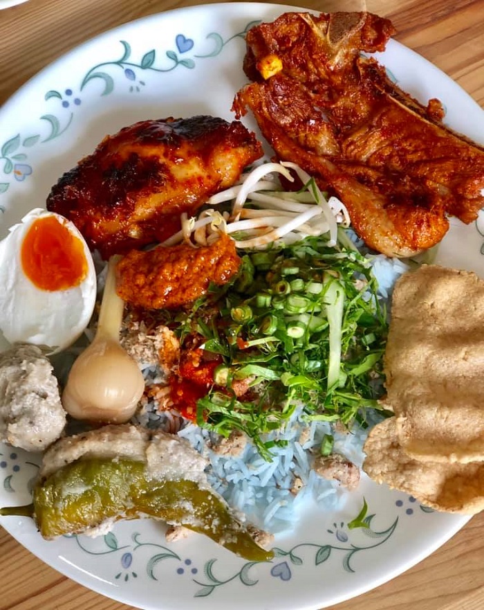 Gambar nasi kerabu Kelantan berwarna biru bersama ulam, solok lada dan lauk pauk yang mencukupi.
