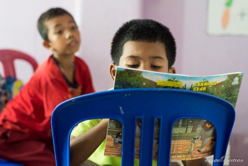Gambar seorang ibu sedang duduk membacakan buku kepada anak perempuannya di rumah.