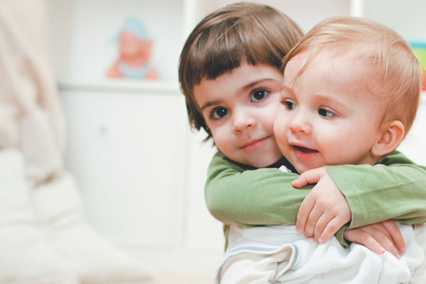 Gambar dua orang adik-beradik kecil sedang duduk berdekatan sambil bermain dan tersenyum.