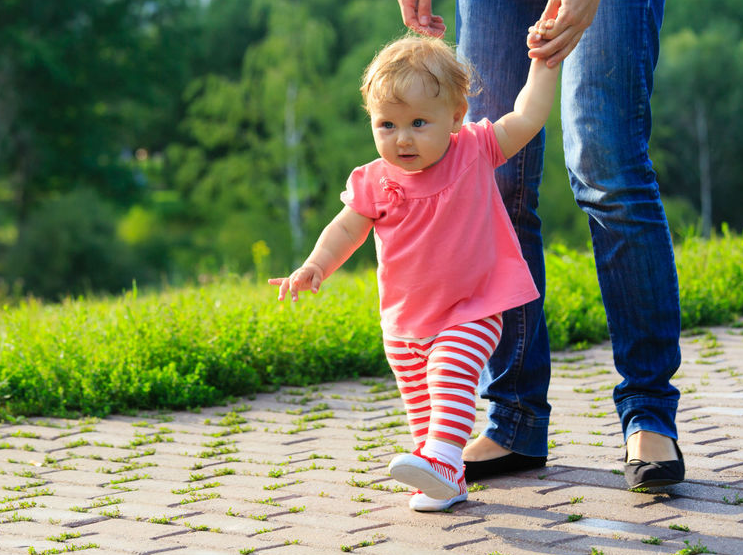 Gambar bayi sedang berlatih berjalan dengan bantuan ibu bapa di dalam rumah.
