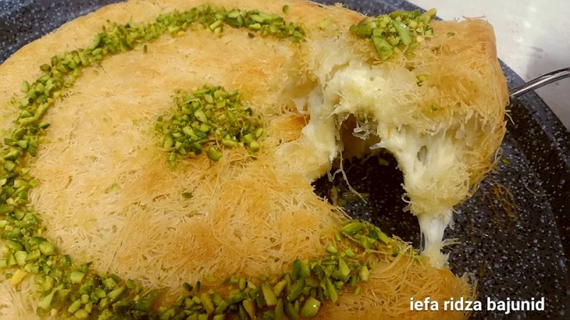 Sekeping Kunafa Nabulsi yang telah dipotong, berwarna keemasan dan dihias dengan kacang pistachio di atasnya.